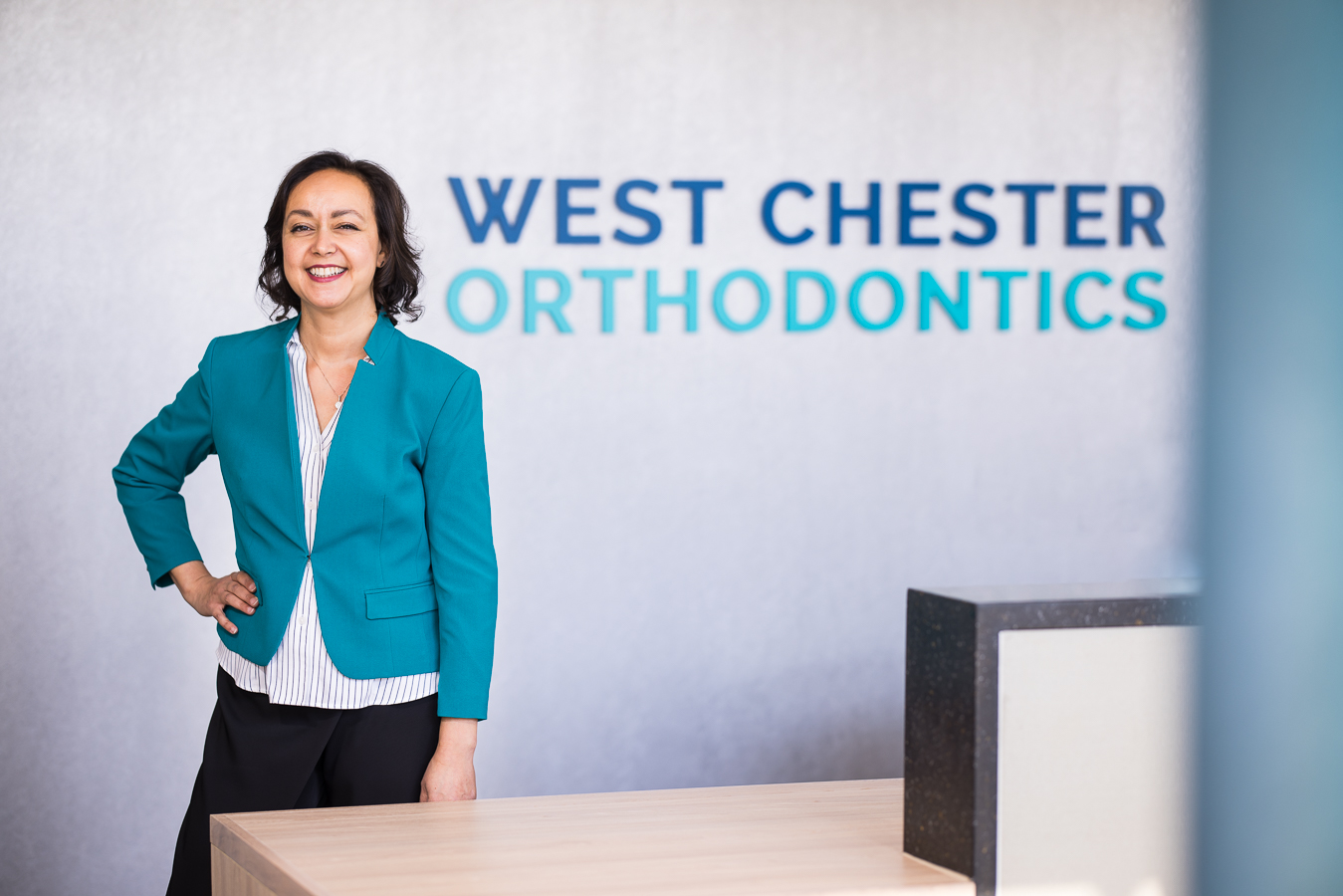 business branding photographer, lisa rhinehart, captures this personalized brand image of dr ferrell owner of west chester orthodontics as she stands by her receptionist desk smiling with pops of teal around her 