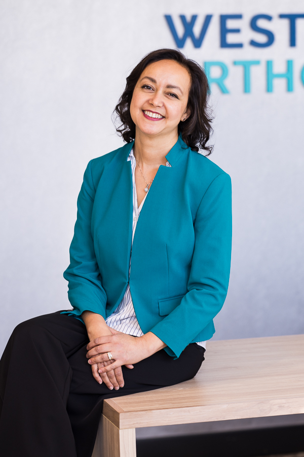 west chester orthodontist, dr ferrell, smiling at the camera as she sits on the receptionist desk in her vibrant teal jacket during her professional headshots for entrepreneurs session of her commercial photography