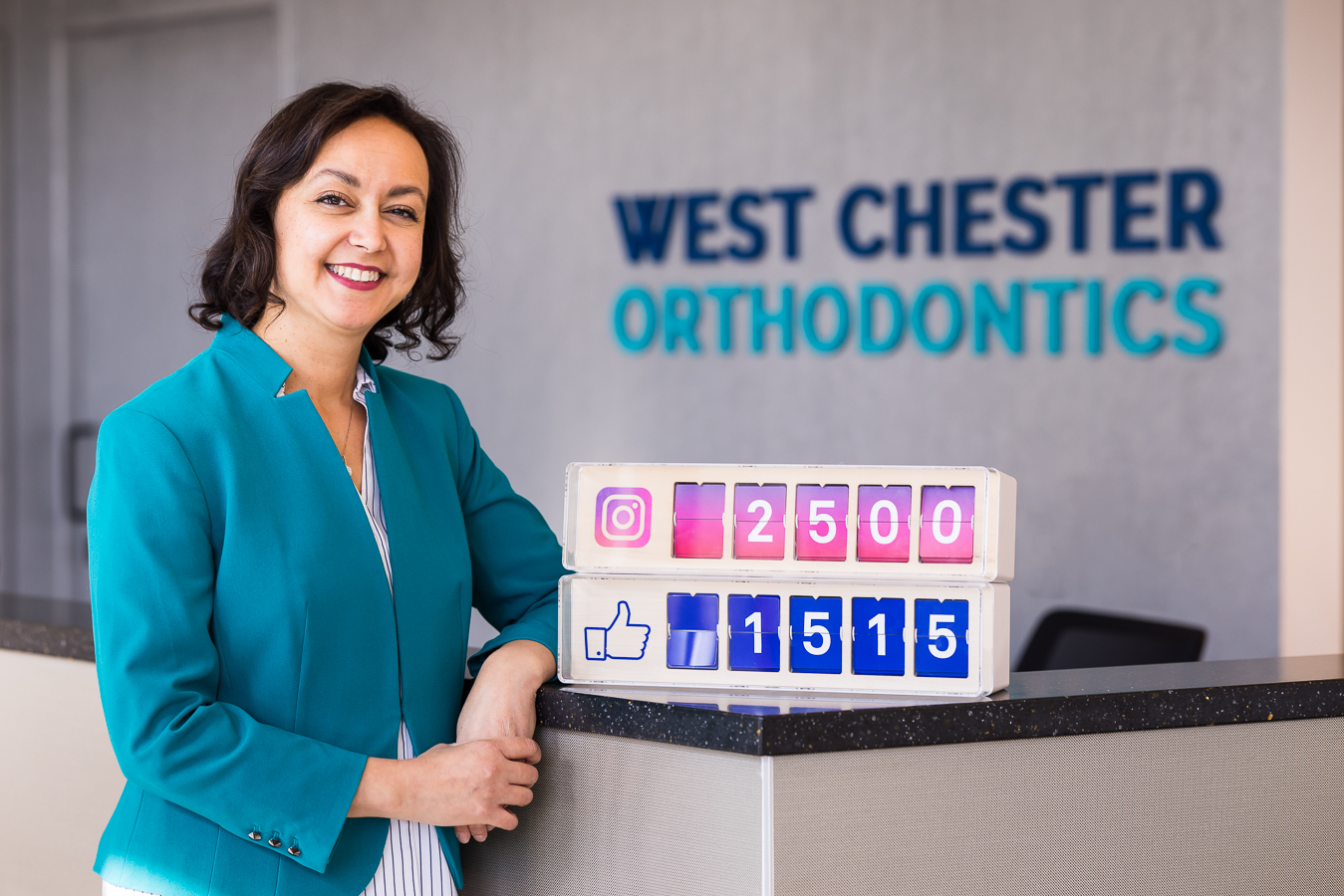 image of dr ferrell from west chester orthodontics as she leans against the receptionist counter in her vibrant teal jacket with her number count as to how many followers they have on Instagram and facebook captured by social media business photographer, lisa rhinehart