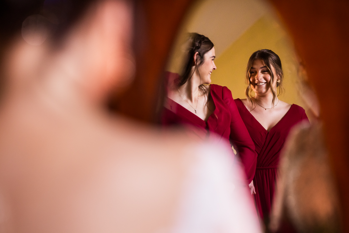 creative wedding photographer, lisa rhinehart, captures this unique perspective of the two bridesmaids in their vibrant deep red dresses through the reflection in the mirror before this outdoor Christmas wedding ceremony in pa