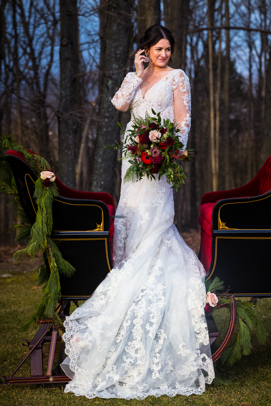 best pa wedding photographer, lisa rhinehart, captures this stunning outdoor bridal portrait of the bride showing off her dress and bouquet while standing up in the sleigh before her outdoor Christmas winter wedding at the stroudsmoor country inn in pa