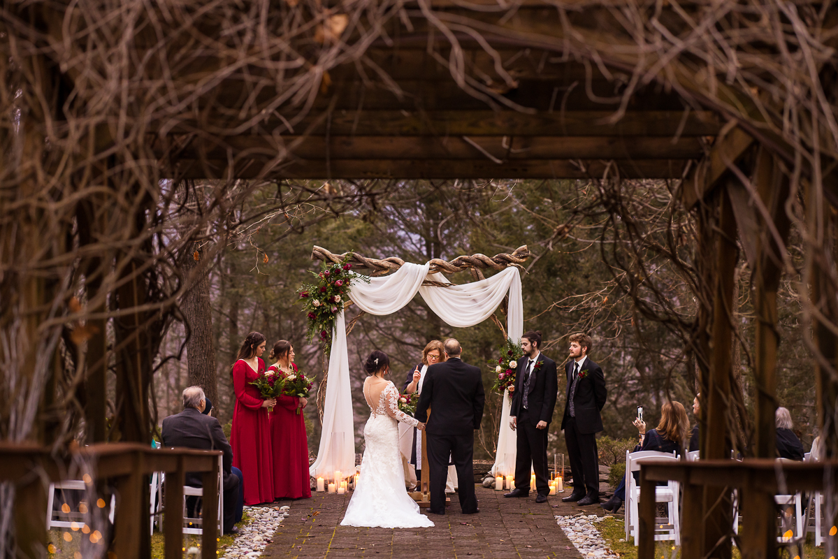 creative stroudsmoor pa wedding photographer, lisa rhinehart, captures this image of the bride and groom as they stand holding hands at the end of the aisle surrounded by cozy wood vibes during their outdoor winter wedding ceremony at auradell 