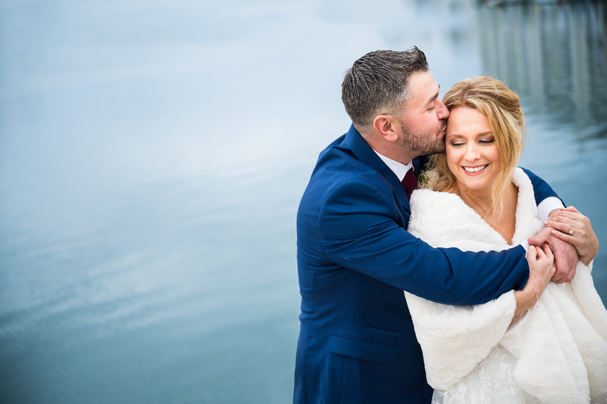 image of the bride and groom as they share a kiss beside the water in New Jersey during their winter first look at their Winter Reeds Stone Harbor Wedding captured by rhinehart photography