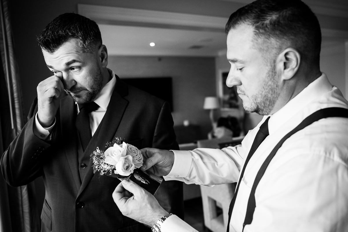 black and white image of the groom as he wipes his tear away as the best man places his boutonniere on which has a photo of his father attached to it at this Christmas beach wedding in New Jersey 