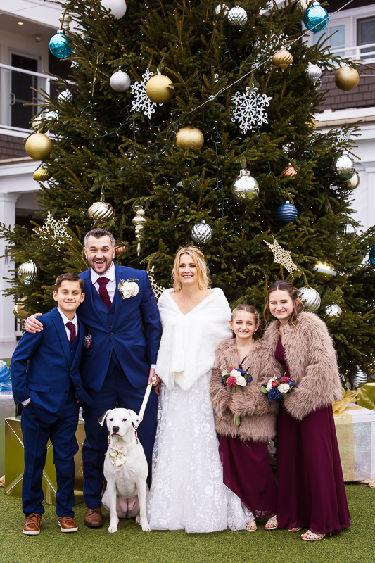 best nj wedding photographer, rhinehart photography, captures this image of the bride, groom, their kids and dog as they stand in front of this giant decorated Christmas tree during their family photos at this Christmas wedding 