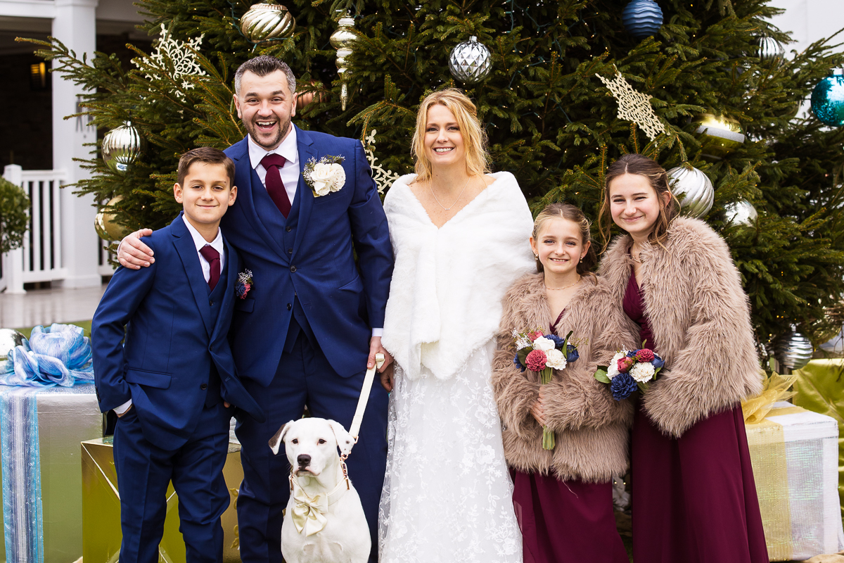 Christmas wedding photographer, rhinehart photography, captures this image of the bride, groom, their kids and their dog as they stand together for their family photo in front of a giant Christmas tree before this winter wedding ceremony 