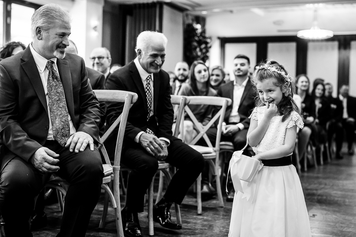 black and white image of the flower girl as she walks down the aisle as guests sit in their chairs and smile as she walks down the aisle during this indoor Christmas wedding ceremony in New Jersey 