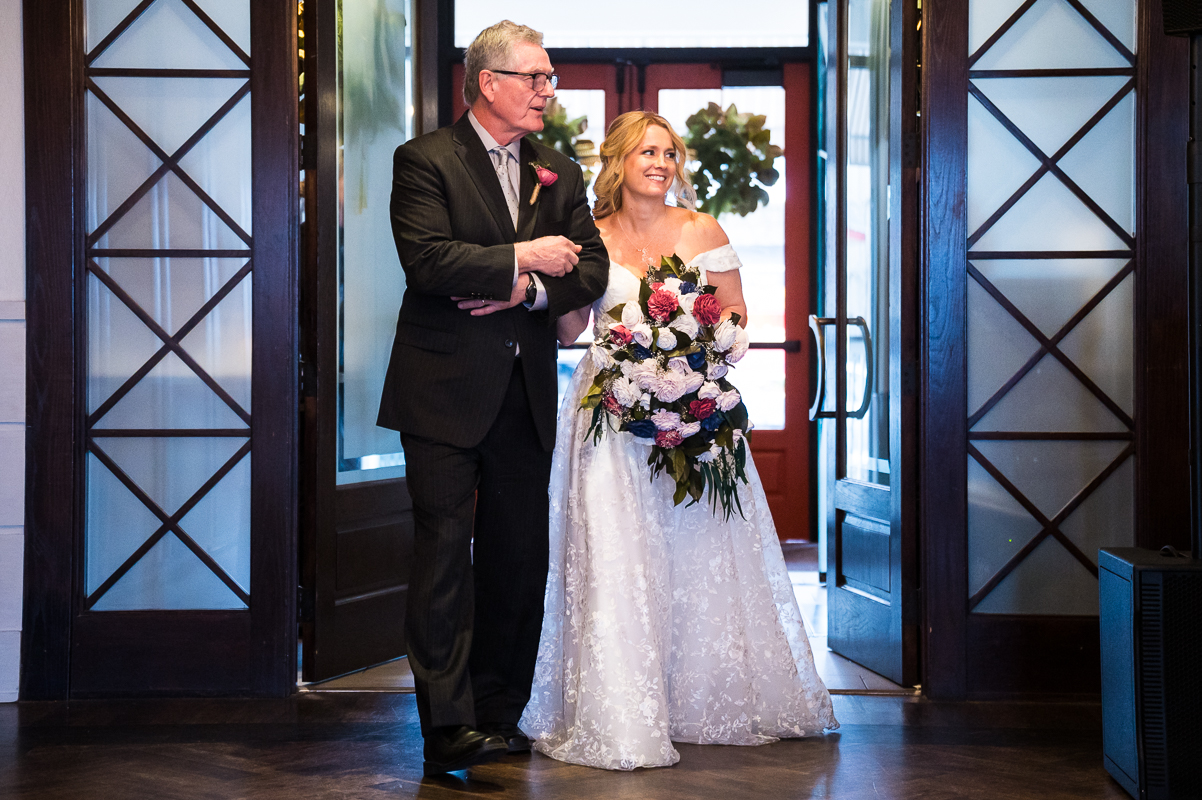 best nj wedding photographer, rhinehart photography, captures this colorful image of the bride and her father as they walk through the door during this indoor winter wedding ceremony held at the reeds in New Jersey 