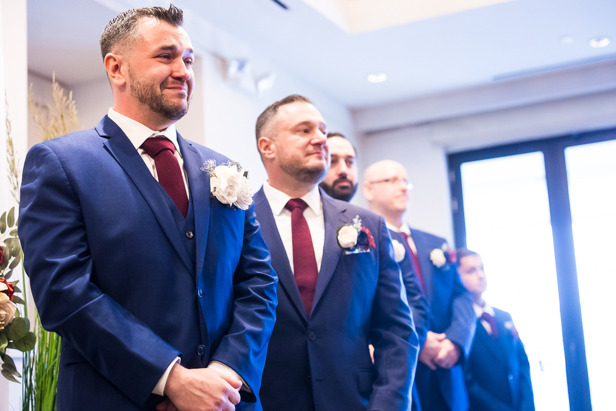 emotional, sentimental image of the groom and his groomsmen in their vibrant blue tuxes as they watch as the bride walks down the aisle during this indoor winter wedding captures by best nj wedding photographer, rhinehart photography 