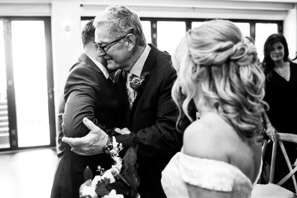 emotional, sentimental black and white image of the groom and the brides father as they share a hug together after walking the bride down the aisle at this Winter Reeds Stone Harbor Wedding