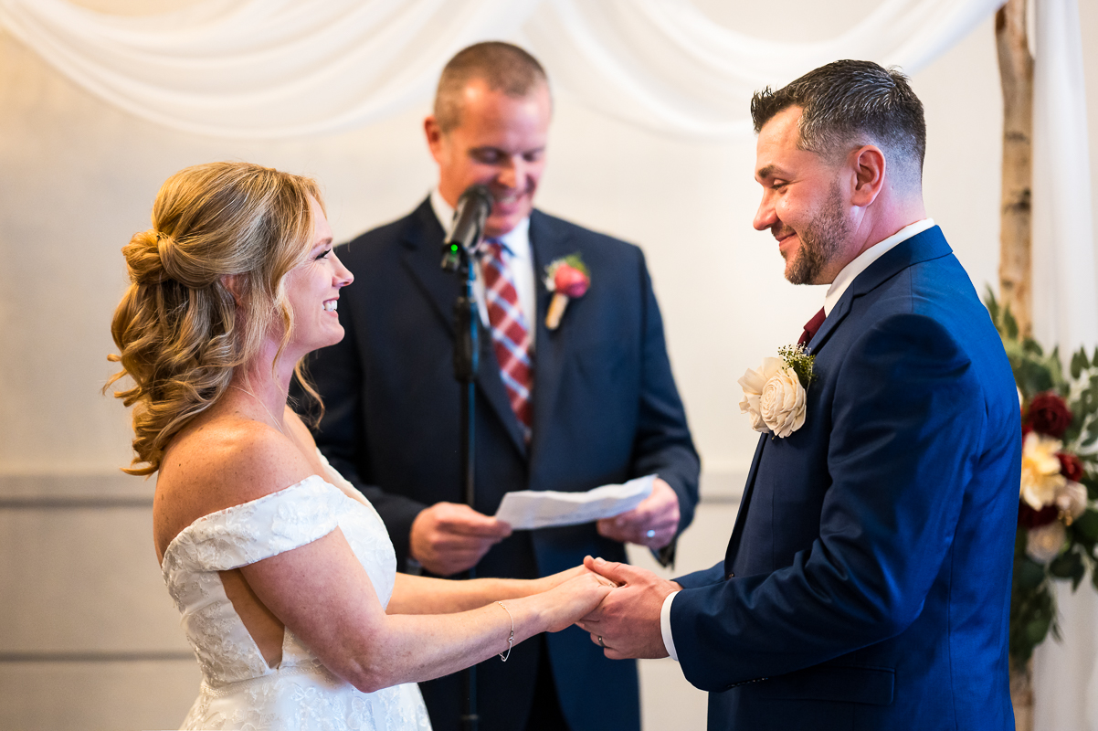 winter wedding photographer, rhinehart photography, captures this image of the bride and groom as they hold hands and smile at one another at the alter during this indoor winter wedding ceremony at the reeds at shelter haven