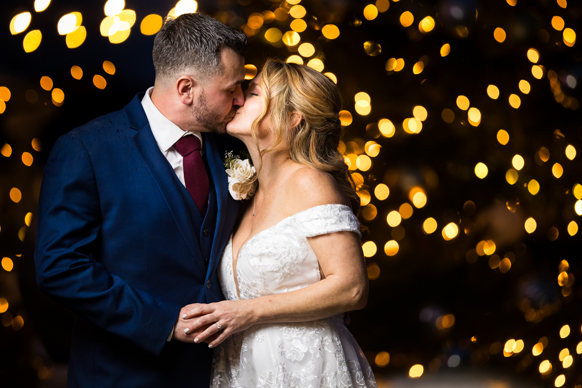 image of the couple as they share a kiss in front of these yellow Christmas lights during this Winter Reeds Stone Harbor Wedding, captured by rhinehart photography