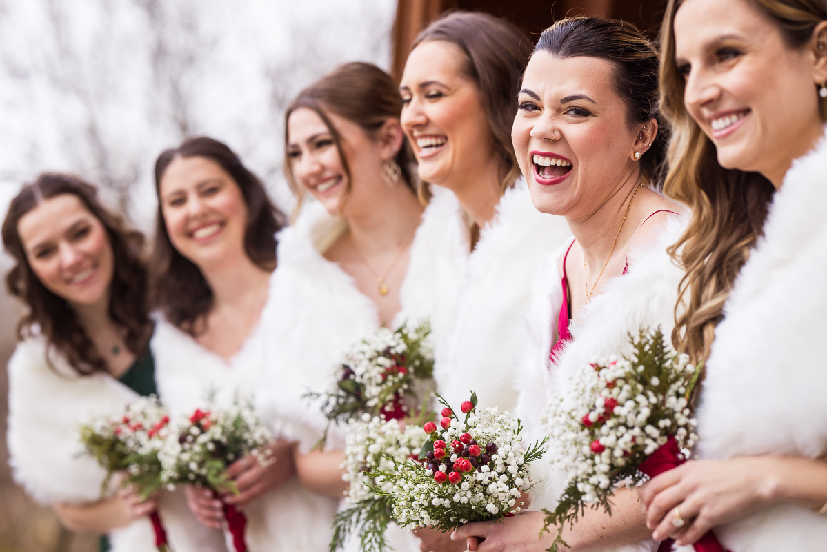 candid moment of the bridesmaids laughing and smiling with one another as they stand with their white shawls covering them and their Christmas themed wedding florals during this outdoor Christmas wedding ceremony in Hallam pa 
