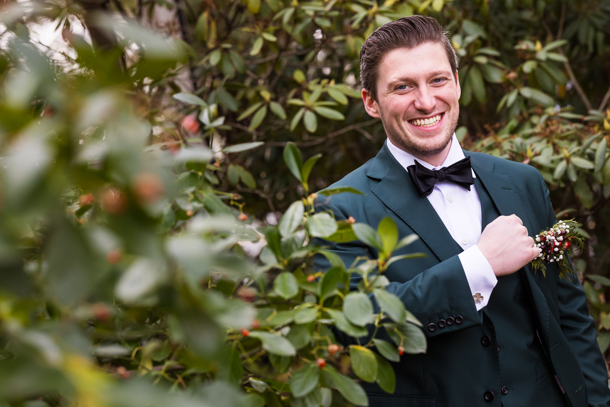 Stone Mill Inn Wedding photographer, Lisa Rhinehart, captures this image of the groom in his green tux surrounded by winter foliage and greenery before this outdoor Christmas wedding ceremony 