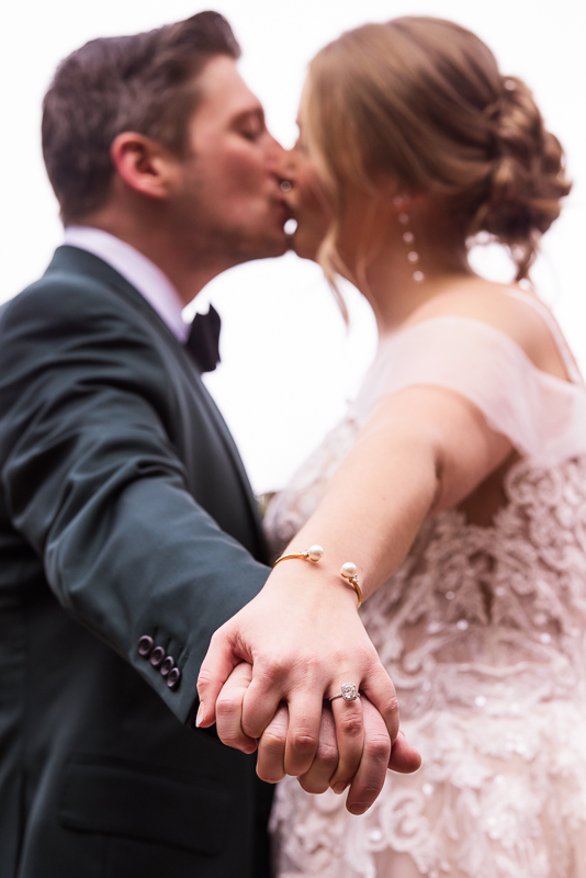 creative wedding photographer, Lisa Rhinehart, captures this unique, creative image of the bride and groom as they kiss in the background while the focus is on their locked hands and rings before their outdoor winter wedding 