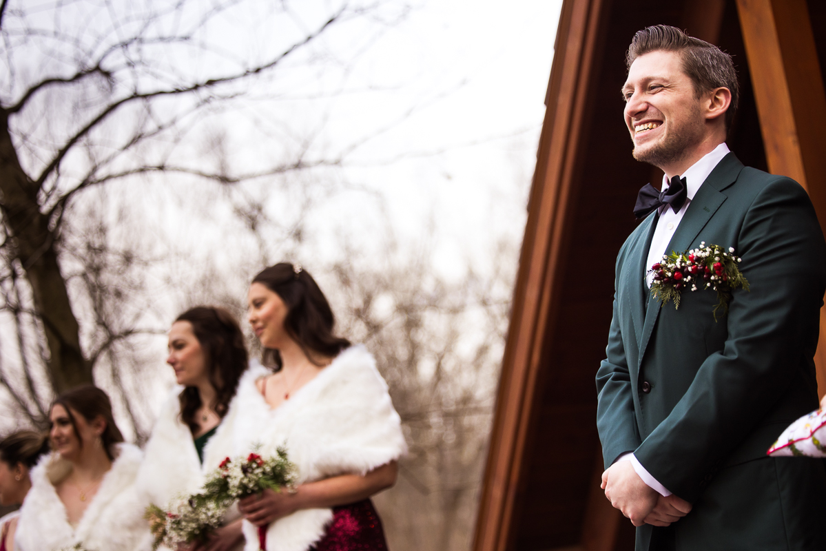 Stone Mill Inn Wedding photographer, Lisa Rhinehart, captures this creative angle of the groom as he beams while watching his bride was down the aisle during their outdoor Christmas wedding ceremony in Hallam pa 