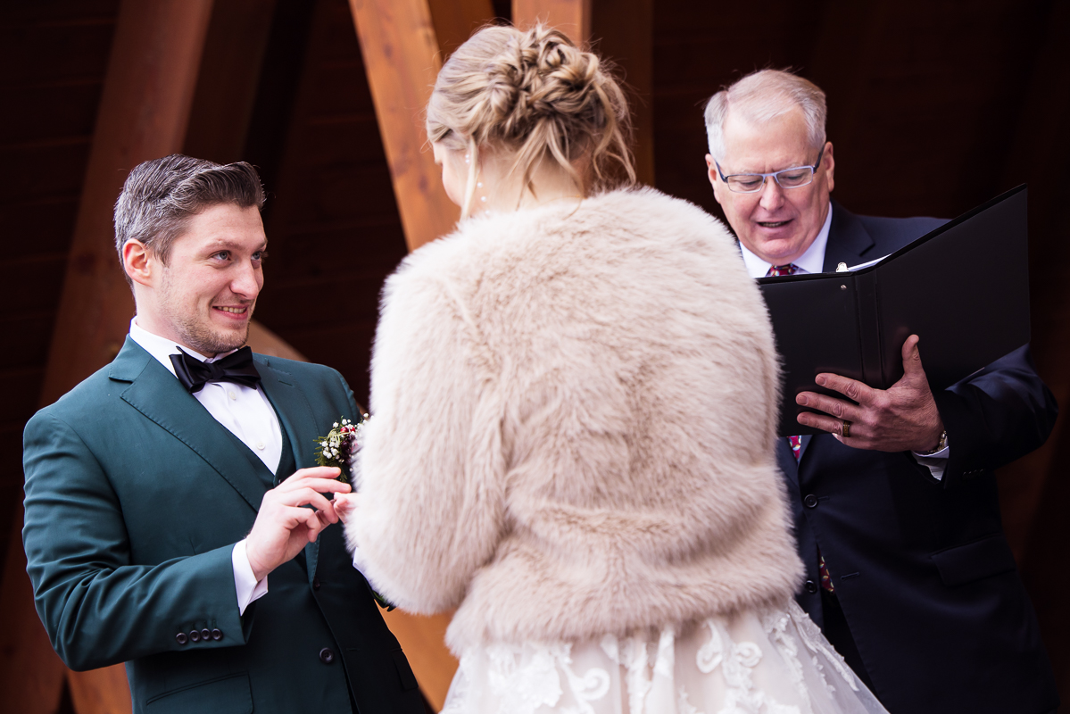 pa wedding photographer, Lisa Rhinehart captures this image of the groom as he smiles while putting the bride's ring on her finger during their outdoor Christmas wedding ceremony at the abbey in Hallam pa 