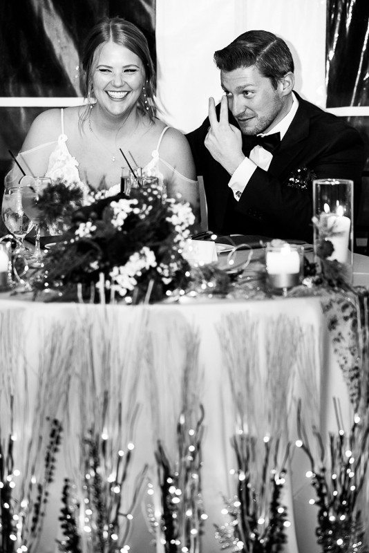 black and white image of the bride laughing and the groom as he makes a motion to a groomsmen giving their speech during the traditions portion of this winter wedding reception in Hallam pa 