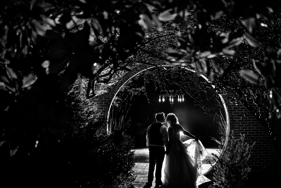 black and white image of the bride and groom as they share a kiss together underneath the moon gate at stone mill inn for their end of the night shots after their Christmas wedding reception 