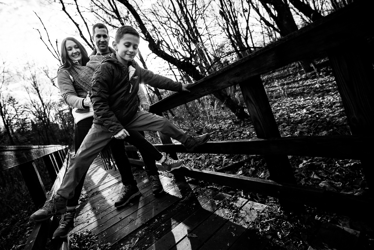 black and white image of the family as one of the boys climbs on the bridge while the rest of the family walks behind him on the wooden bridge at the wildlands conservancy in Emmaus pa with family photographer, Lisa Rhinehart 