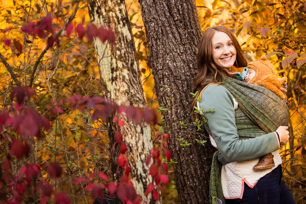 vibrant, colorful image of the mom with her youngest child as she smiles at the camera while leaning against the tree surrounded by vibrant, colorful red and yellow leaves during this outdoor fall family session at the wildlands conservancy in emmaus pa 