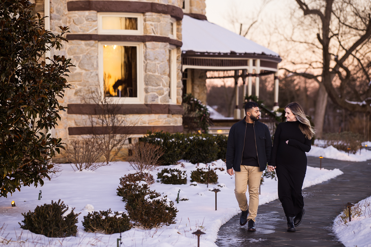 candid, traditional portrait of the husband and wife as they hold hands and walk on the pathway outside of the willows at ashcombe mansion during this winter maternity session in central pa 
