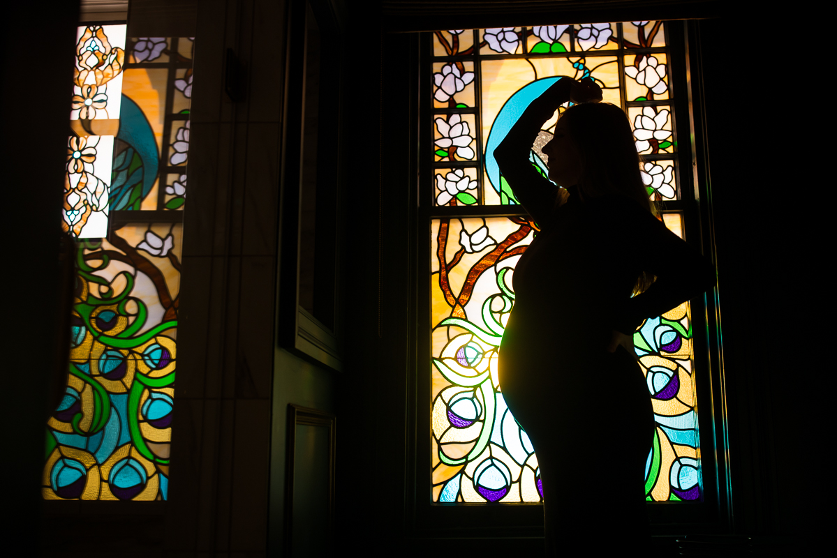 Creative Mechanicsburg family photographer, Lisa Rhinehart, captures this creative, unique silhouetted portrait of the wife as she poses in front of this vibrant, colorful stained glass window during this indoor maternity session at the willows at ashcombe mansion in central pa 