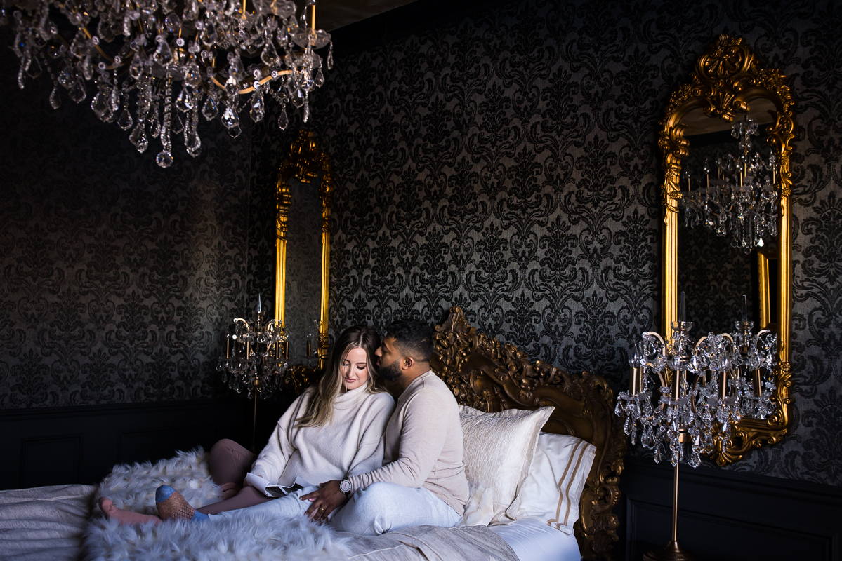 best pa maternity photographer, Lisa Rhinehart, captures this cozy portrait of the husband and wife as they sit together on the bed surrounded by crystal chandeliers during their maternity session at the willows at ashcombe mansion in central pa 