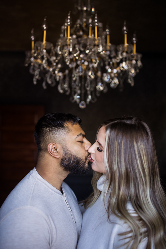 image of the husband and wife as they share a kiss together underneath the chandelier inside of the willows at ashcombe mansion in central pa during this cozy maternity session 