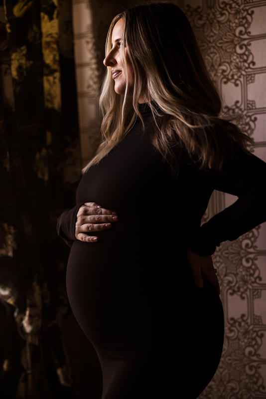 stunning dark and moody maternity portrait of the mom as she poses in her black body con dress while holding her belly during this cozy maternity session at the ashcombe mansion captured by Lisa Rhinehart 