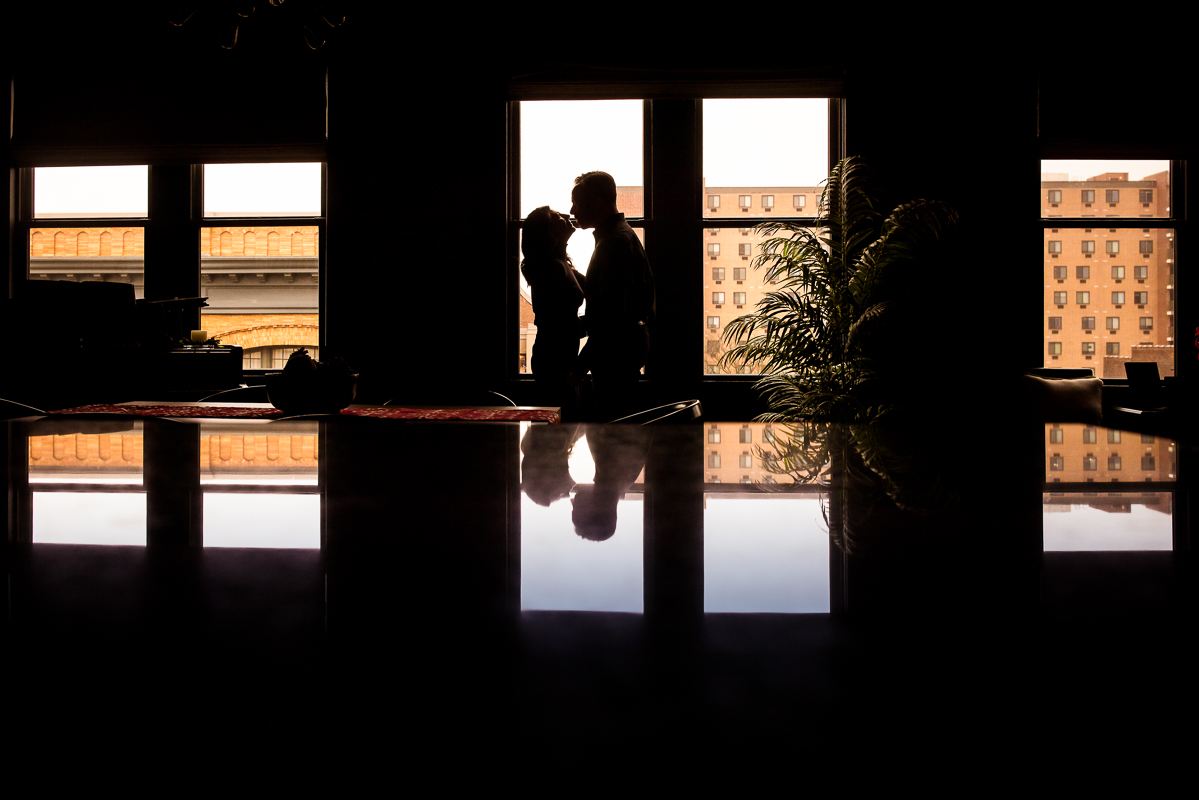creative Yorktowne Hotel Wedding engagement photographer, Rhinehart Photography, captures this creative, reflective image of the couple as they stand in front of the window during this York engagement session inside of their home at the lofts on george in york pennsylvania 