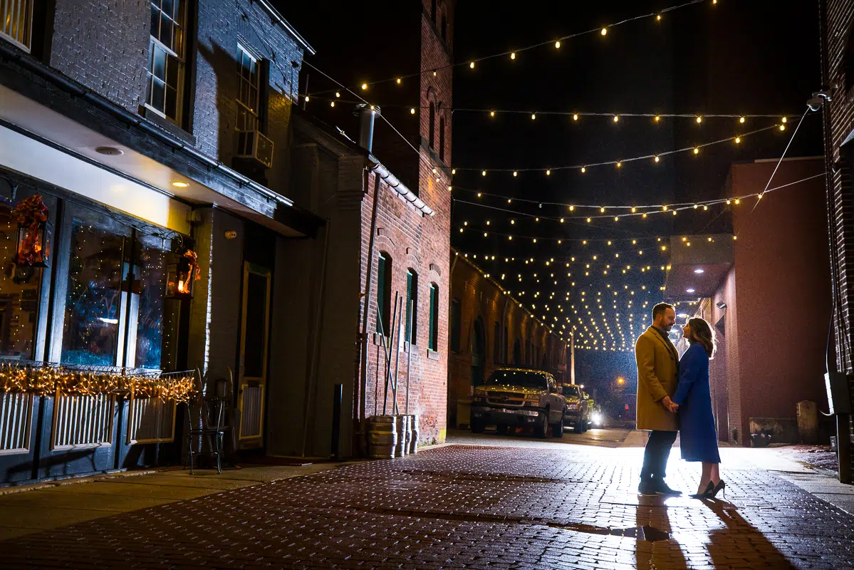 Yorktowne Hotel Wedding engagement photographer, Lisa Rhinehart, captures this night time romantic portrait of the couple as they stand together on this red brick road with twinkle lights above their head during their outdoor winter engagement session in york pa