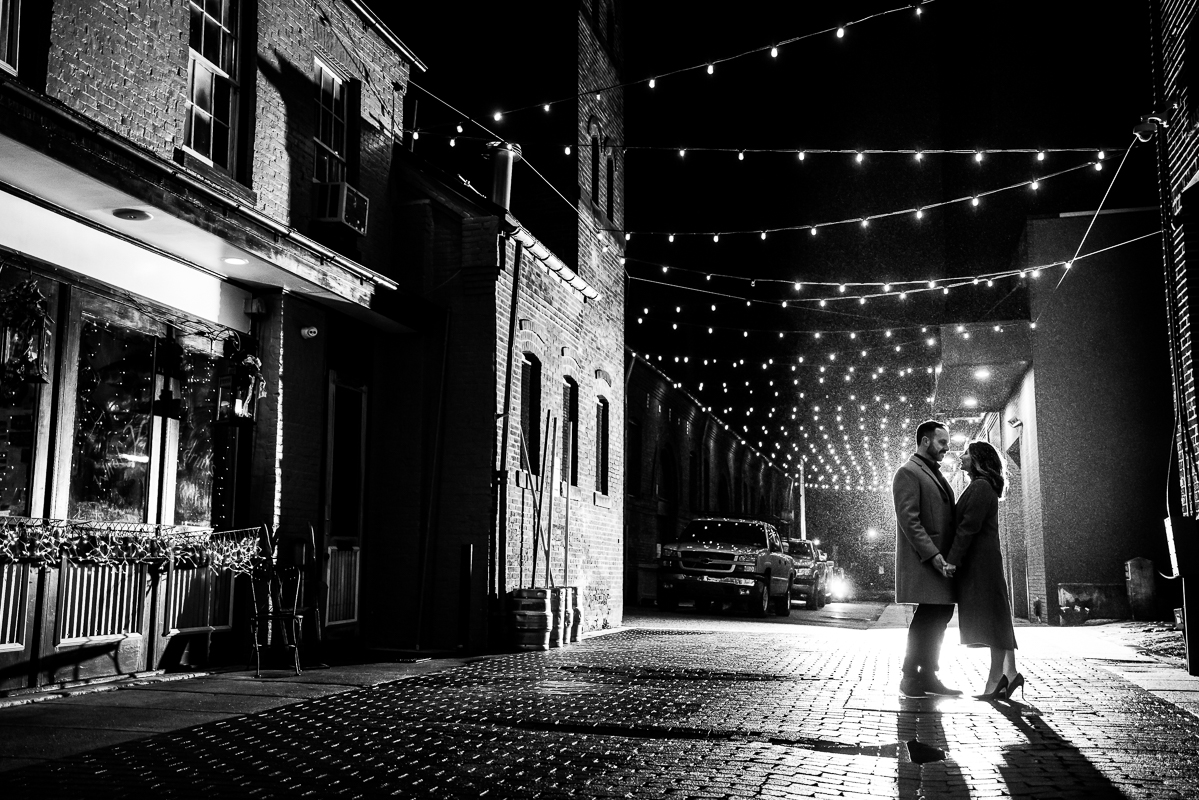 Yorktowne Hotel Wedding engagement photographer, Lisa Rhinehart, captures this night time romantic portrait of the couple as they stand together on this red brick road with twinkle lights above their head during their outdoor winter engagement session in york pa 