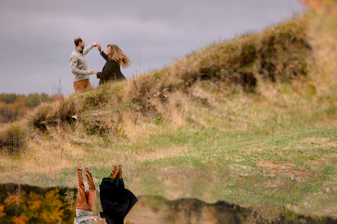 A couple dances on a grassy hill, their reflections shimmering in the water below. Set outdoors with autumn foliage and a cloudy sky, this scene is perfectly captured by the best engagement photographer.